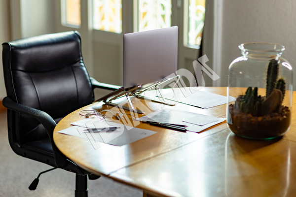 Home workstation for smart working. In the image wooden table, laptop, glasses, greasy plant in glass vase, papers and pens, desk chair and window