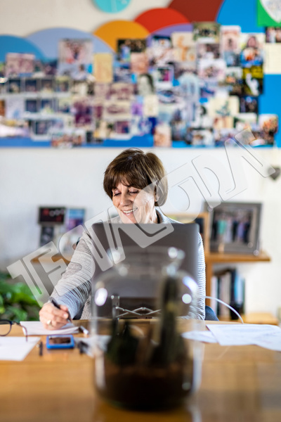 Smiling Senior female professional takes notes with pen and paper working remotely from home in a video meeting with her laptop on a wooden table