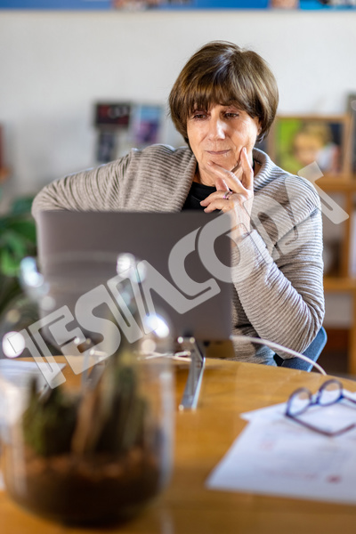 Aged lady Senior Consultant with hand on cheek working remotely from home in a video meeting with her laptop on a wooden table with papers and glasses