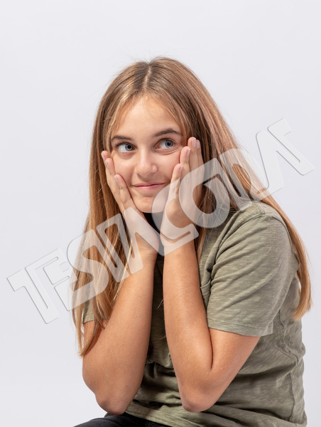 Cute, cunning, smiling teenager girl in green t-shirt with blond long hair face-in-hands looks to the side with sly, amazed, smart and funny expression