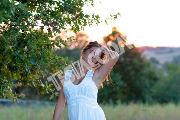 A romantic young girl with blond long hair, dressed in white, poses at sunset near the magic tree in countryside